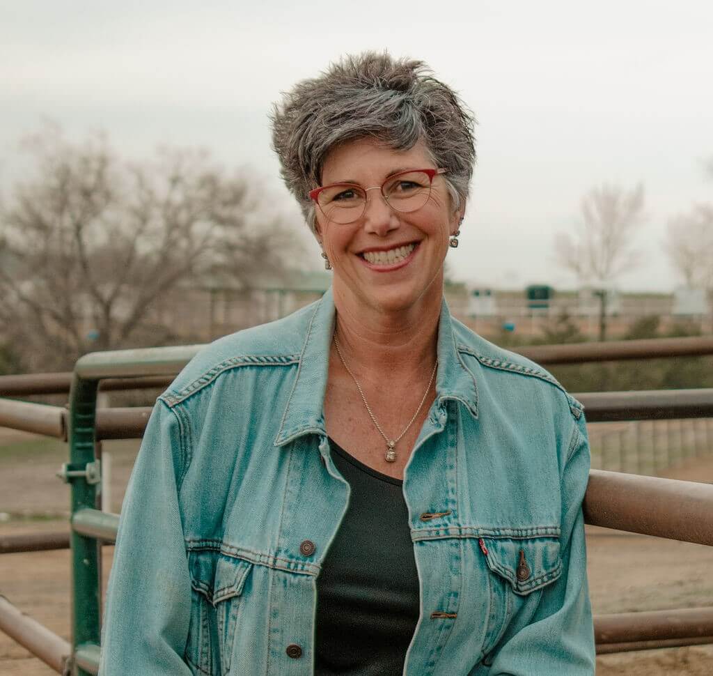 A woman with short gray hair and glasses smiles while standing in front of a metal fence, wearing a denim jacket and black top, with a blurred outdoor background.