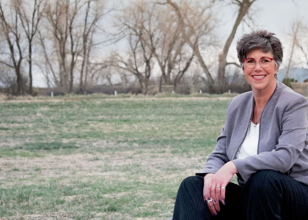 A person with short gray hair and glasses sits outdoors on grass, wearing a gray blazer and white shirt. Leafless trees are in the background.