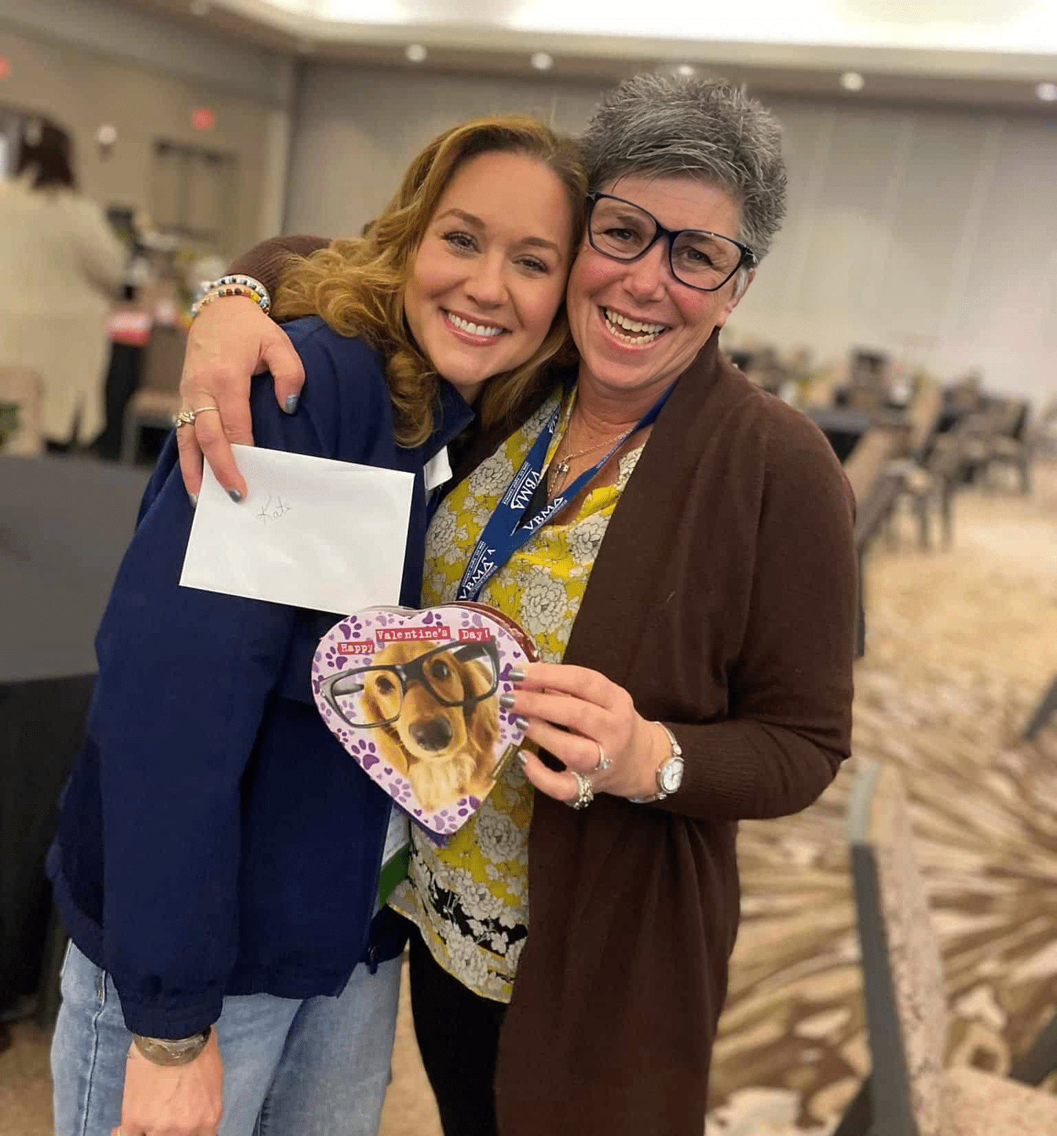 Two women smiling and hugging indoors; one holds an envelope, the other holds a heart-shaped item with a dog wearing glasses printed on it.