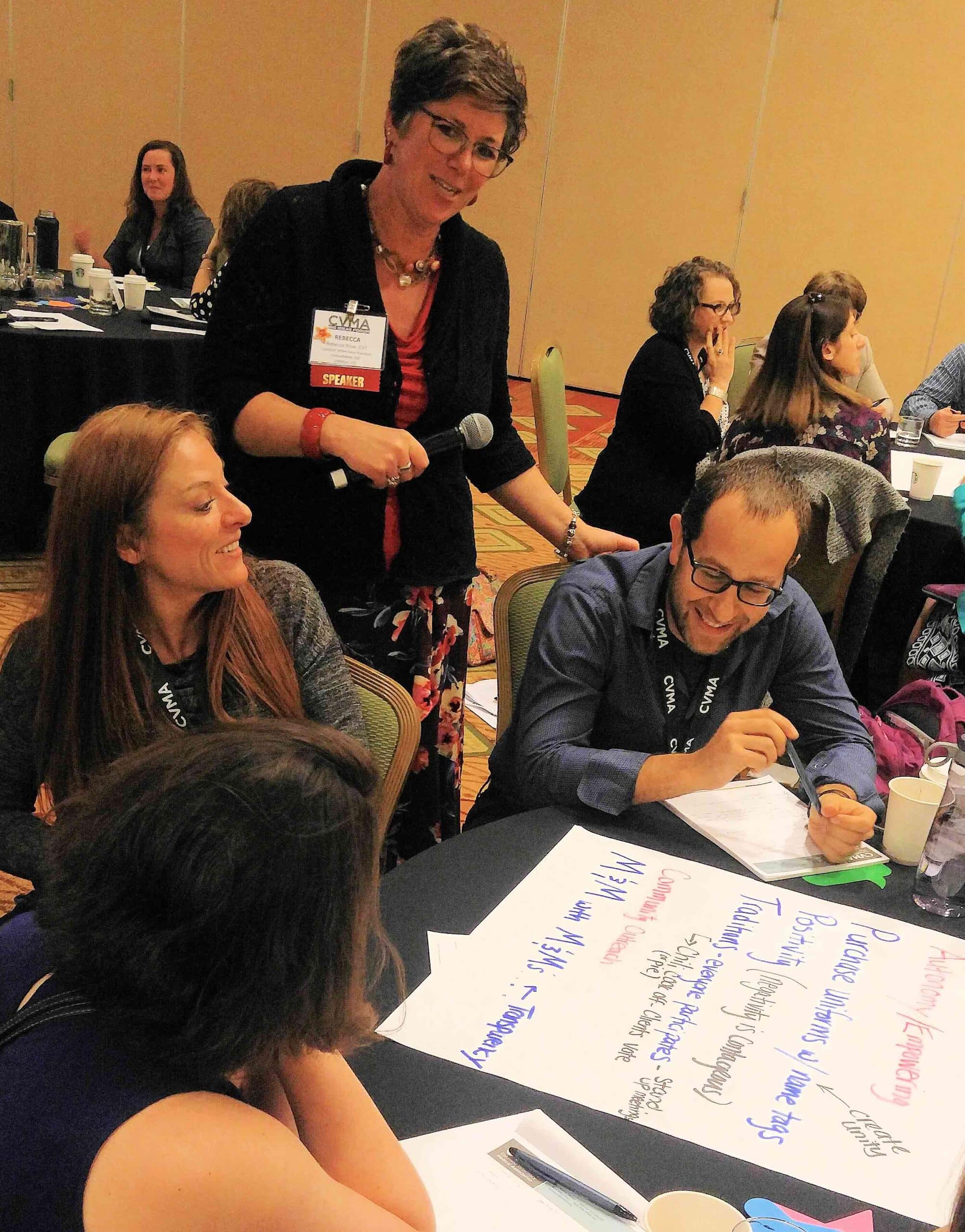 A woman with a microphone stands beside three seated people who are discussing and writing notes on a large sheet of paper at a conference.