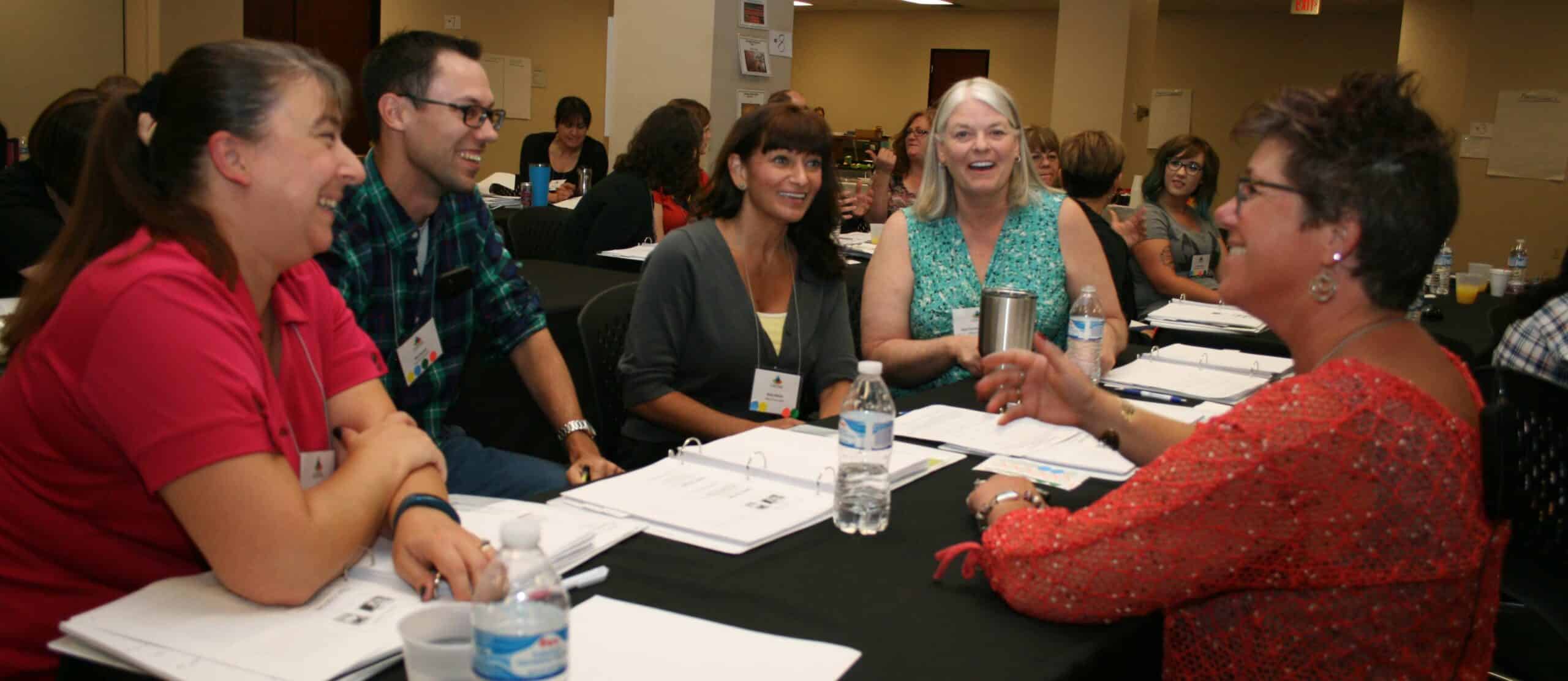 Five adults sit around a table, smiling and talking during a meeting or workshop. Papers, notebooks, and water bottles are on the table. Other groups are visible in the background.