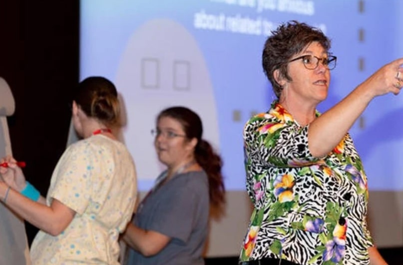 A woman in a colorful shirt gestures while two others in scrubs write on a board, with a projected presentation in the background.