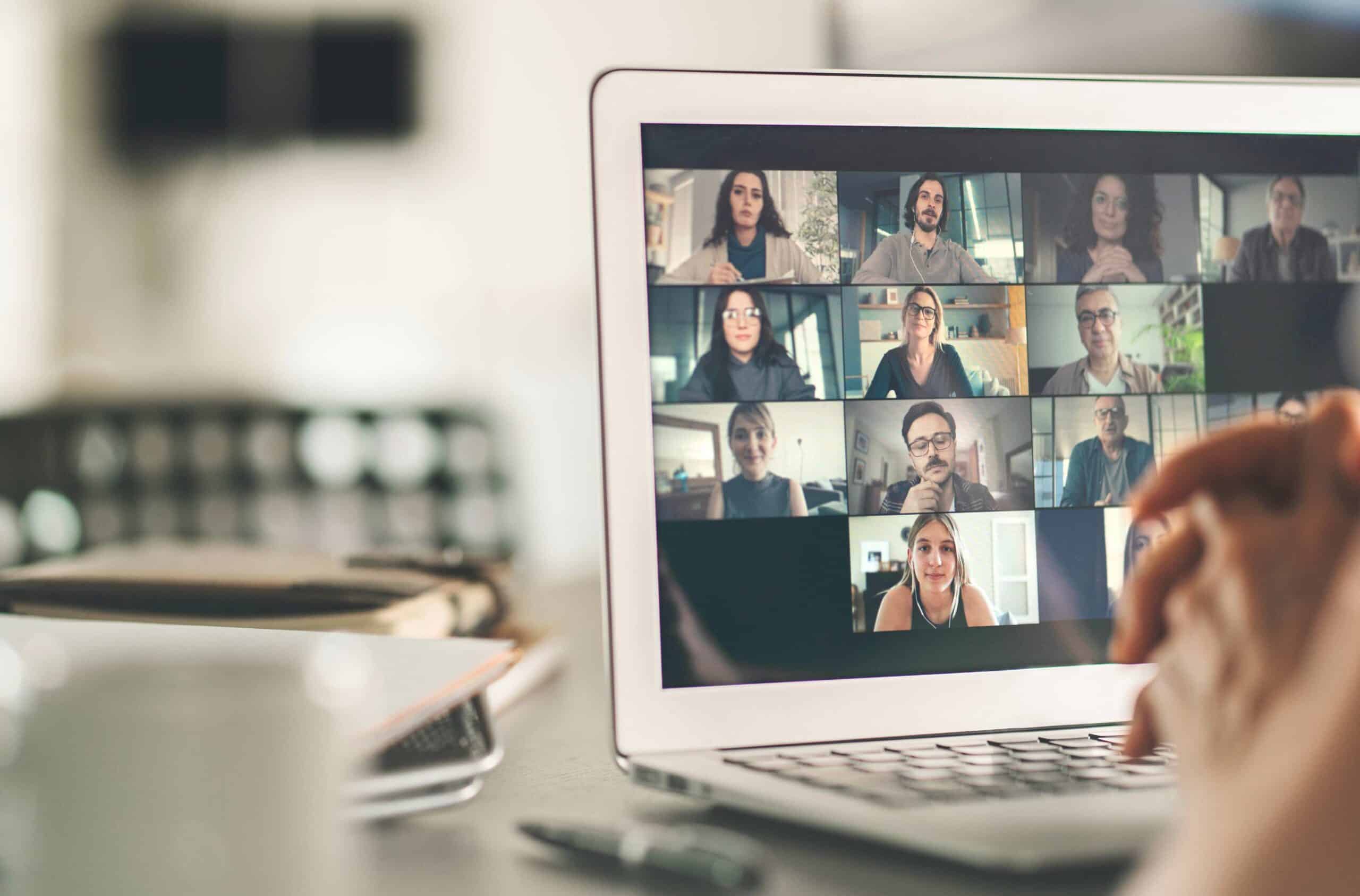 Laptop screen showing a video conference with twelve participants in individual video boxes; hands are visible in the foreground.