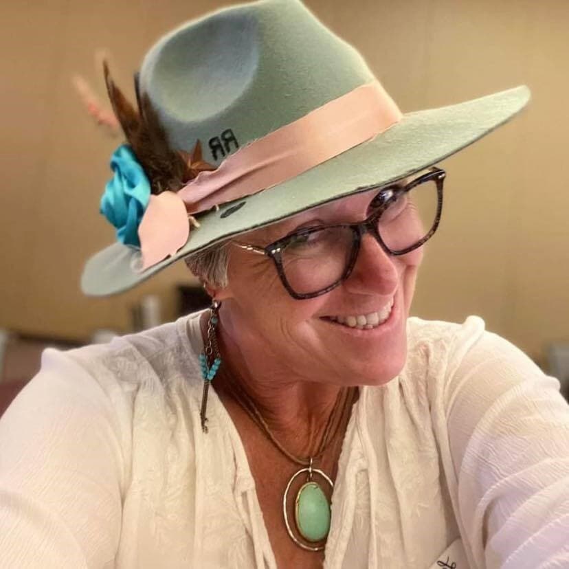 A person wearing glasses and a green hat with a pink ribbon and turquoise flower, smiling at the camera. They have a white shirt and a turquoise pendant necklace.